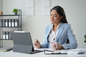 Smiling businesswoman working remotely and drinking coffee, analyzing financial charts and reports on a digital tablet