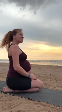 Young pregnant active woman doing exersice outdoors, stratching her body and meditate on the sea, beach at sunset in the evening