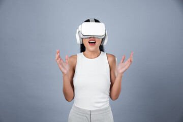 Smart female standing with gray background wearing VR headset connecting metaverse, futuristic cyberspace community technology. Elegant woman excited seeing generated virtual scenery. Hallucination.