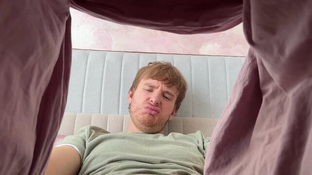 Young man having urological problems and bad potency, looking under the blanket in bed in bedroom