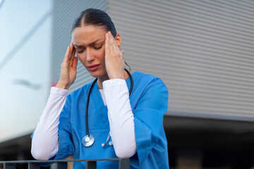 Female healthcare worker in blue scrubs feeling stressed, touching temples with closed eyes while standing outside hospital