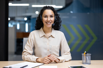 Smiling female customer service representative wearing headset in modern office workspace. Confident professional offering assistance, working with documents and tablet desk corporate environment.