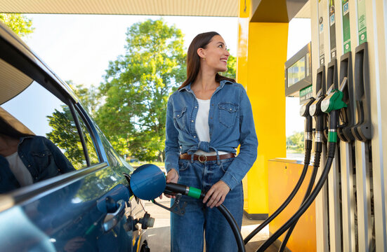 Happy woman refueling her car at petrol station, cheerful female driver standing near vehicle holding filling gun nozzle and looking at screen