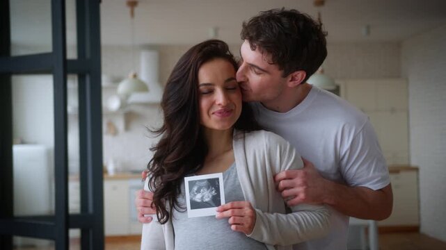 Happy pregnant couple pregnancy medical insurance health parents smiling laughing home man guy male father kiss woman girl female mother in cheek together holding showing USD picture ultrasound photo