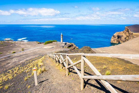 Wooden trail fence and lighthouse building at Capelinhos peninsula with volcanic beach and ocean in background, Faial island, Azores, Portugal
