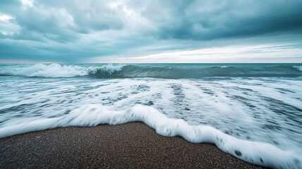 A serene view of waves crashing onto a sandy beach under a cloudy sky.
