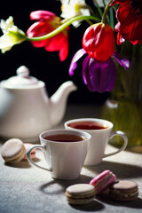 Teapot with two cups of tea, macarons, and tulips on a table with a shadowy background
