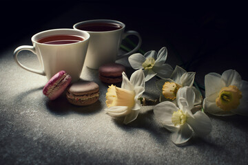Two cups of tea with macarons and white flowers on a dark background, creating a cozy and elegant still life scene