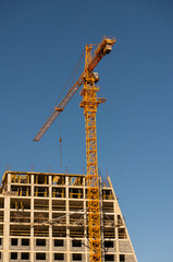 Towering yellow crane at a construction site against a clear blue sky, with a building under construction and visible scaffolding © lappoa