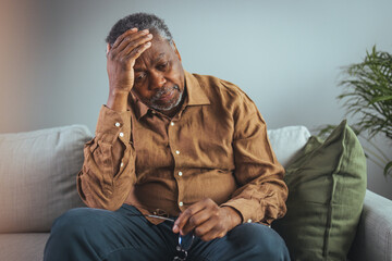 Elderly Black Man Experiencing Discomfort on Couch. A senior black male grips his glasses, showing...