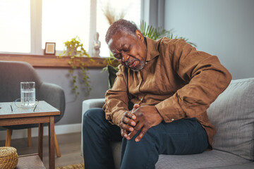 A senior African American man grips his knee in discomfort while seated on a gray sofa, his face...