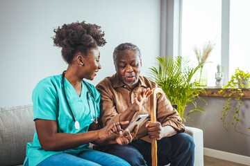 A mid-adult Black nurse in scrubs attentively assists an elderly Black man with his medication,...