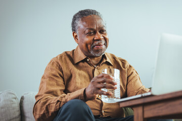 A senior black man in casual attire is seated comfortably on a sofa, engaging with a laptop and holding a glass of water.
