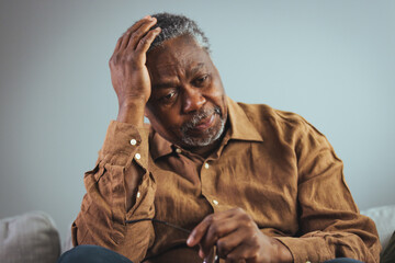 A senior black male looks distressed while seated on a sofa, his hand on his head suggesting a...