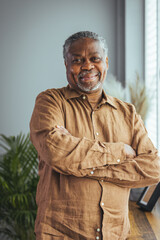 A cheerful senior Black male stands in a home setting, arms crossed, wearing a casual brown shirt,...