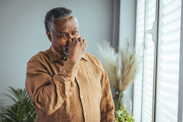 A pensive senior Black gentleman stands indoors, dressed in casual attire, hand resting...