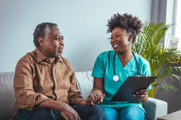 A black mid-adult nurse takes notes while engaging with an elderly male patient in a home setting,...