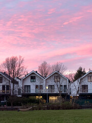 Row of charming houses with modern architecture at sunset, featuring gabled roofs and large windows, set against a serene evening sky in a suburban area.