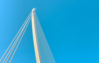 Low-angle view of a modern suspension bridge against a clear blue sky, emphasizing structural engineering, sleek design, and urban architecture.