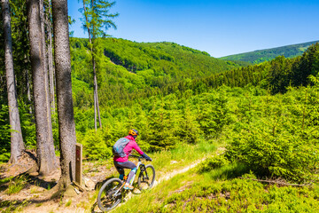 Woman with backpack riding mountain bike on single track trail among green hills in Jizera...
