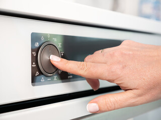 Woman adjusting modern white oven in kitchen.  Female hand pushing and turning knobs on oven control panel