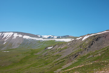 Fototapeta premium Vivid layered landscape with sunlit steep green rocky mountain wall with snow cornice against high snowy mountain range under clear blue sky. Glacier on top in bright sun. Sunny day in high mountains.