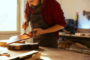Craftsperson in carpentry shop starting restoration project on wood block, applying lacquer on surface. Carpenter coloring plank with paintbrush, doing handwork in studio