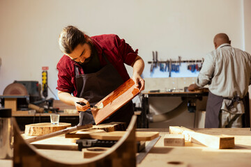 Carpenter brushing varnish on wood to build up protective layer with african american coworker in background. Woodworker lacquering plank after sanding surface next to BIPOC apprentice working