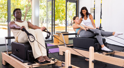 African-american man and young european woman exercising with pilates reformers. Latin woman instructor assisting them with workout.