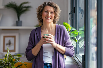 Mature woman drinking a cup of coffee while looking at camera standing next to the window at home.