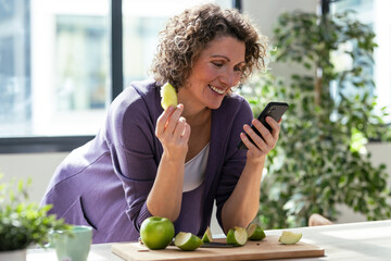 Pretty mature woman eating an apple while using smartphone in the kitchen at home