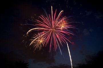 A large red and blue firework and two smaller yellow gold fireworks at dusk against a partially cloudy night sky. Copy space.