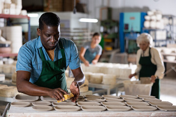 Skillful african-american man ceramist in apron polishing new plates in workshop.