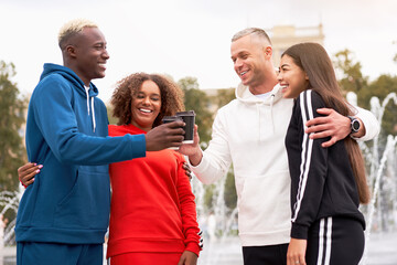 Multi-ethnic friends in sportswear toasting coffee cups. African-American Asian Caucasian student spending time together. Multiracial friendship. Happy smiling couples in park.