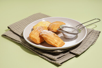 Plate with delicious madeleines, sieve and powdered sugar on green background