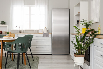 Interior of light kitchen with stylish fridge, counters, dining table and houseplant