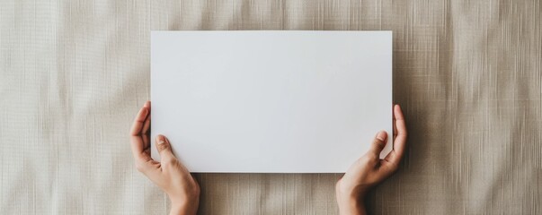 Hands holding blank white sheet on beige fabric background, top view. Minimalism and simplicity concept