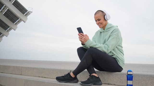 Mature sports woman smiling listening to music with headphones and using her smartphone to watch a workout app, resting after a cardio workout. Athletic female let up after running training