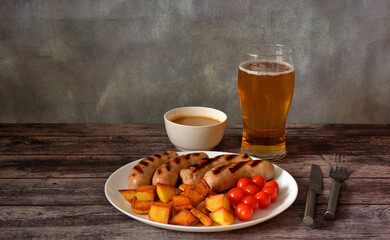 A glass of light beer, next to a plate of grilled sausages with fried potatoes, tomatoes and mustard sauce on a wooden table.