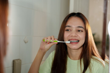 Asian teenage girl brushing teeth with braces in bathroom. Long straight brown hair, light green ribbed top. Indoor setting with tiled walls. Focused on dental hygiene and morning routine.