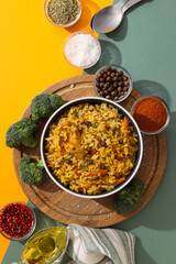 Pilaf in bowl, board and spices on yellow and green background, top view