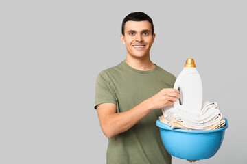 Young man with clean towels in basin and bottle of detergent on grey background