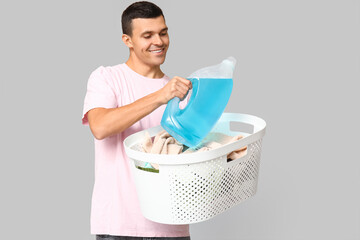 Young man with laundry basket and bottle of detergent on grey background