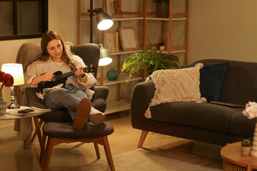 Obraz premium Beautiful young woman sitting in armchair and playing ukulele in evening