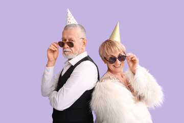 Mature couple in party hats with sunglasses on lilac background