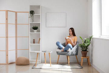 Young woman having cup of tea while sitting in armchair in living room