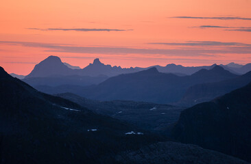 Midnight sun in arctic summer landscape of Norway. Silhouettes of mountain ranges in sunset colours of blue, orange and purple. Few clouds on clear dawn and dusk sky. Moody mountain range landscape