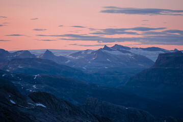 Midnight sun in arctic summer landscape of Norway. Silhouettes of mountain ranges in sunset colours of blue, orange and purple. Few clouds on clear dawn and dusk sky. Moody mountain range landscape