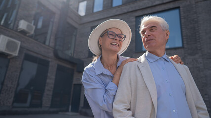 A woman in a hat and glasses hugs an elderly man in a white jacket from behind. Romantic relationships of mature people. 