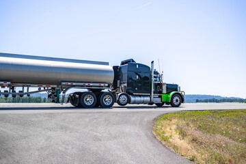 Black classic big rig semi truck tractor with green fenders transporting liquid cargo in tank semi...
